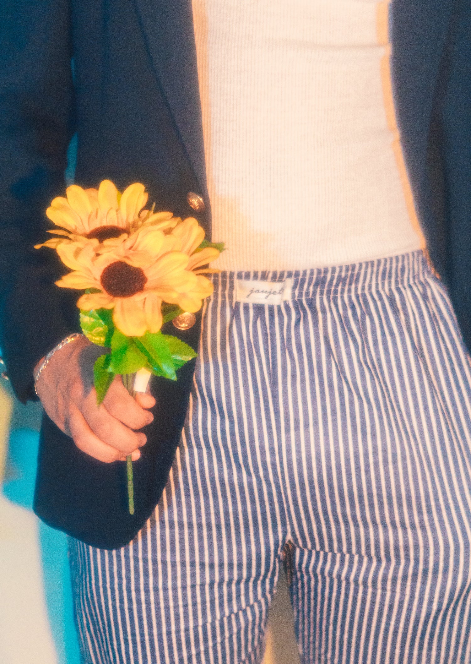 Male wearing pinstriped joujete pants, and a t-shirt and blazer, while holding a few sunflowers.